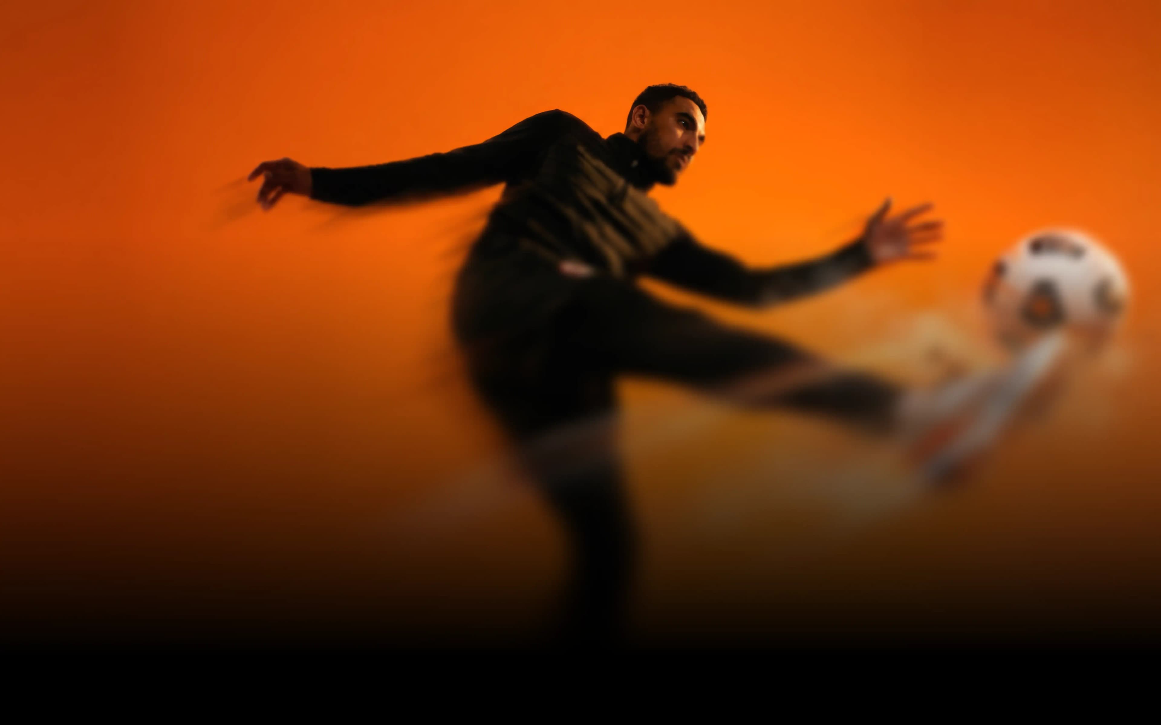 Male soccer player in dark attire powerfully kicks a soccer ball against an orange background.