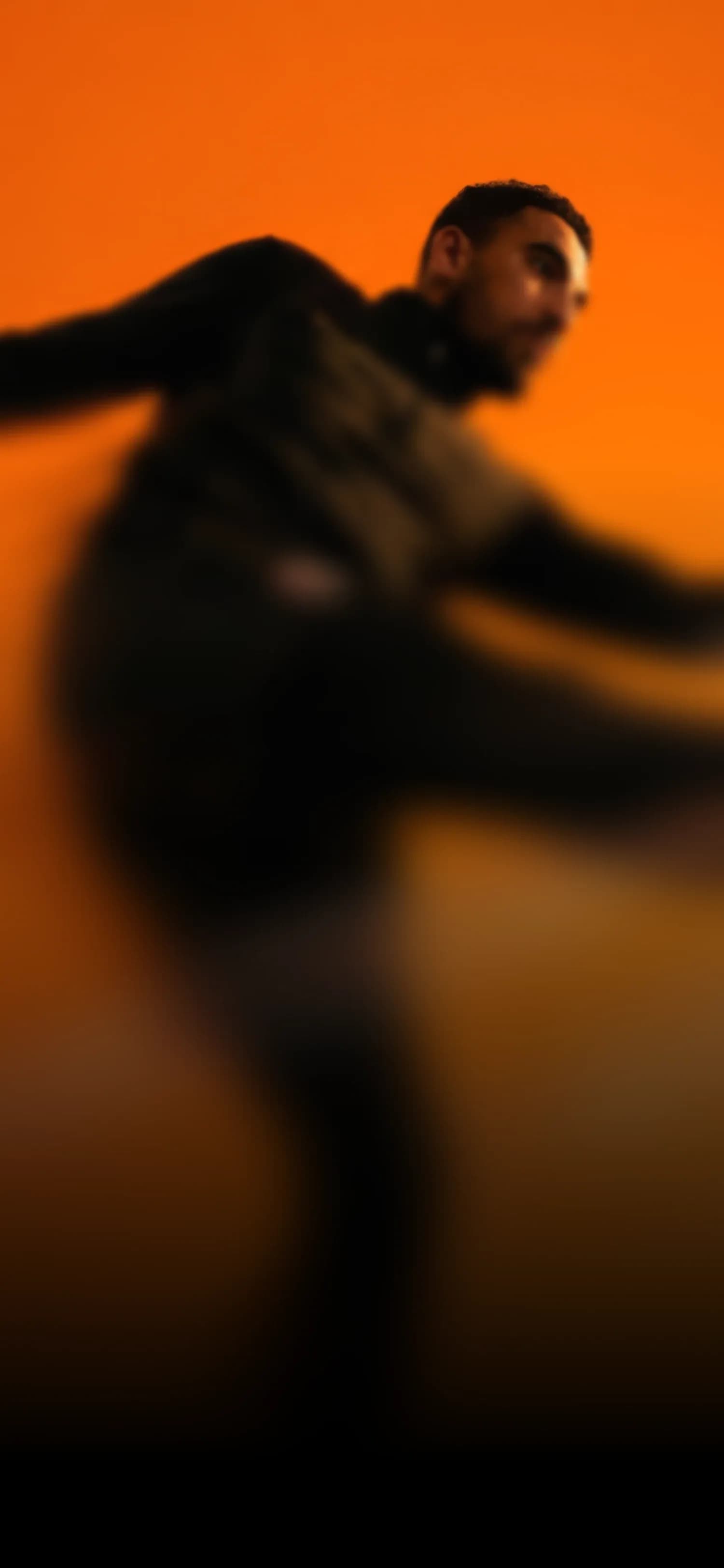 A person in dark clothing appears to be dancing or moving energetically against an orange background.